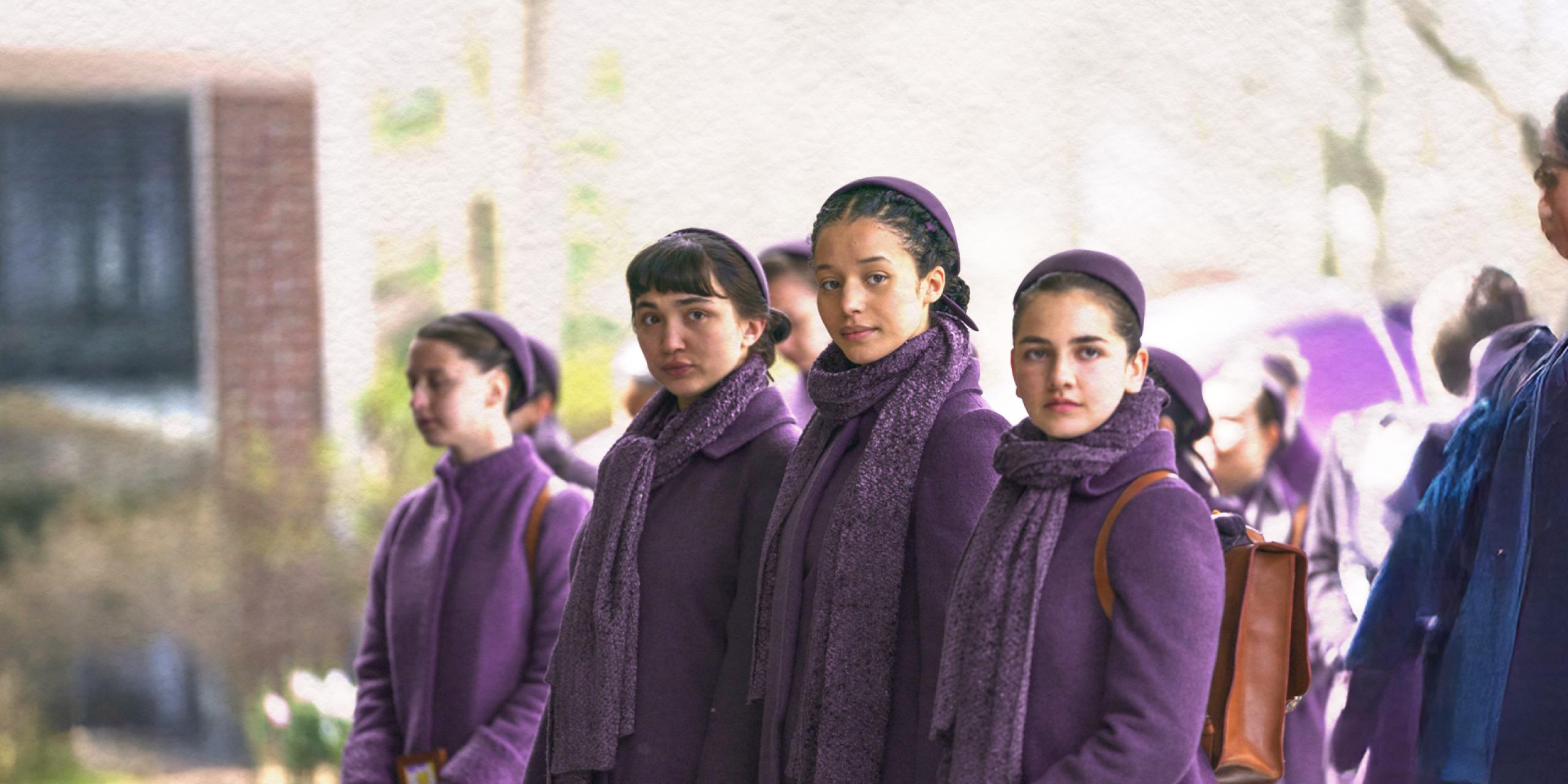 Four characters from The Testaments TV show stand in a line. They are wearing purple and outisde.