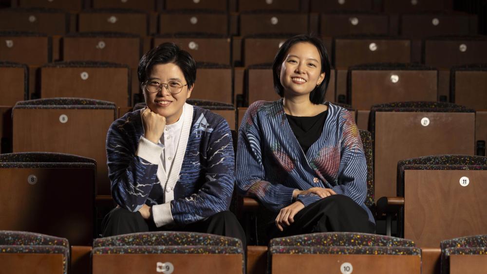 Two individuals sit in an auditorium. One has short hair and glasses. They have their head resting on their fist. The other sits straight and has longer hair. Both are wearing cardigans and smiling at the camera.